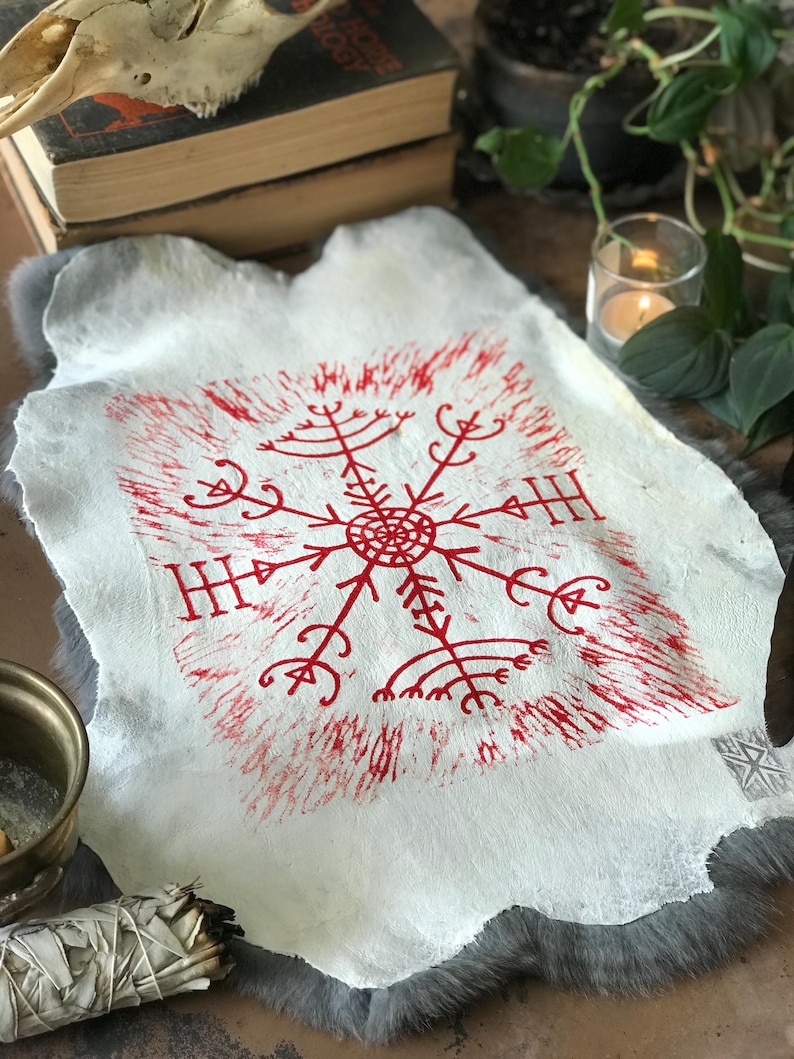 May include: A white, textured hide with a red, stylized snowflake design. The artwork features a central circular element with radiating lines and symbols. The hide is displayed on a gray, furry surface, with other items in the background.