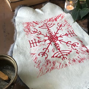 May include: A white leather hide with a red, stylized snowflake design. The design is centered on the hide and has a textured, hand-printed appearance. The hide is displayed on a gray fur-like surface.