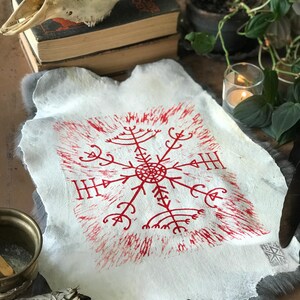 May include: A white, textured piece of paper with a red, symmetrical symbol at the center. The symbol has a circular center with radiating lines and arrow-like shapes. The paper is set on a gray, furry surface, with a skull and books in the background.