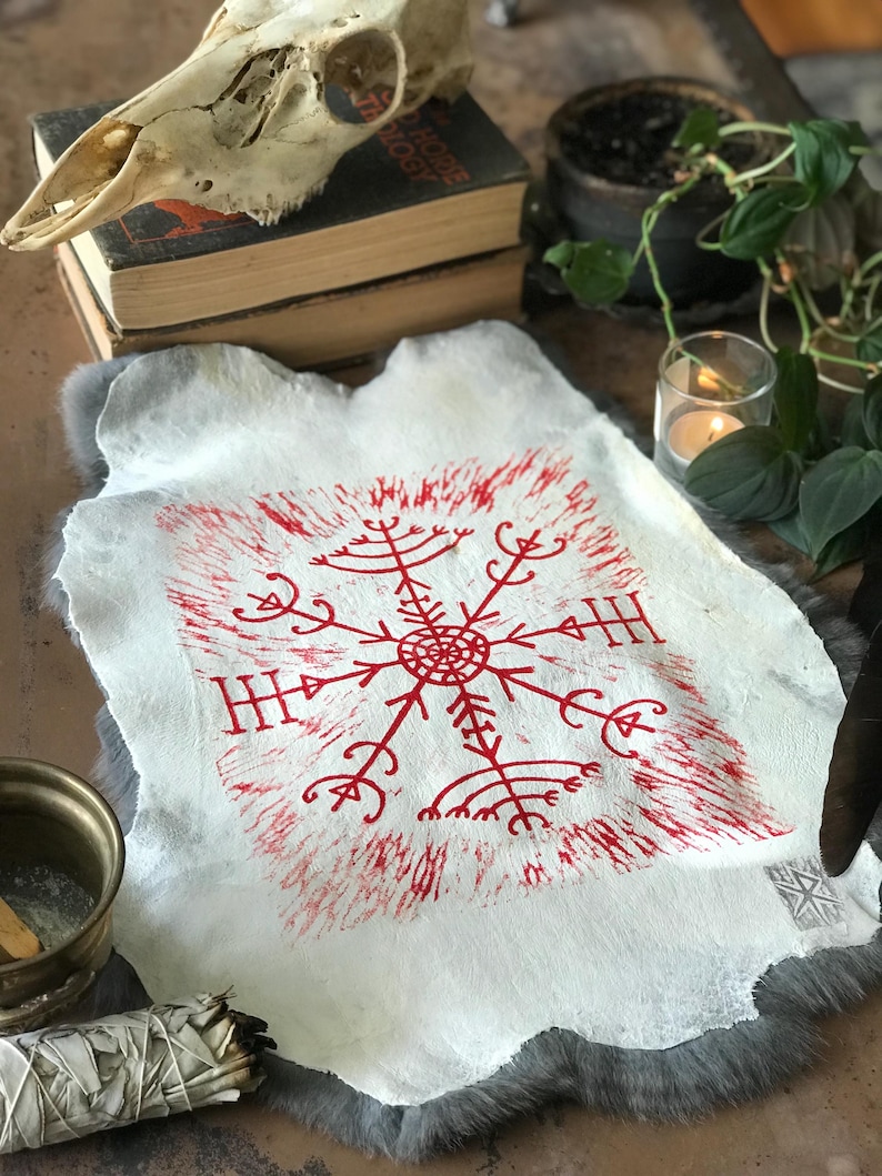 May include: A white, textured piece of paper with a red, symmetrical symbol at the center. The symbol has a circular center with radiating lines and arrow-like shapes. The paper is set on a gray, furry surface, with a skull and books in the background.
