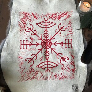 May include: A white leather hide with a red, stylized, symmetrical design in the center. The design is a series of lines and shapes radiating from a central circle. The hide is on a gray, furry surface.