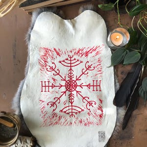 May include: A white, irregularly shaped piece of material with a red, stylized symbol at the center. The symbol is surrounded by a red, textured border. The item is displayed on a gray, furry surface, with a candle and other decorative items in the background.