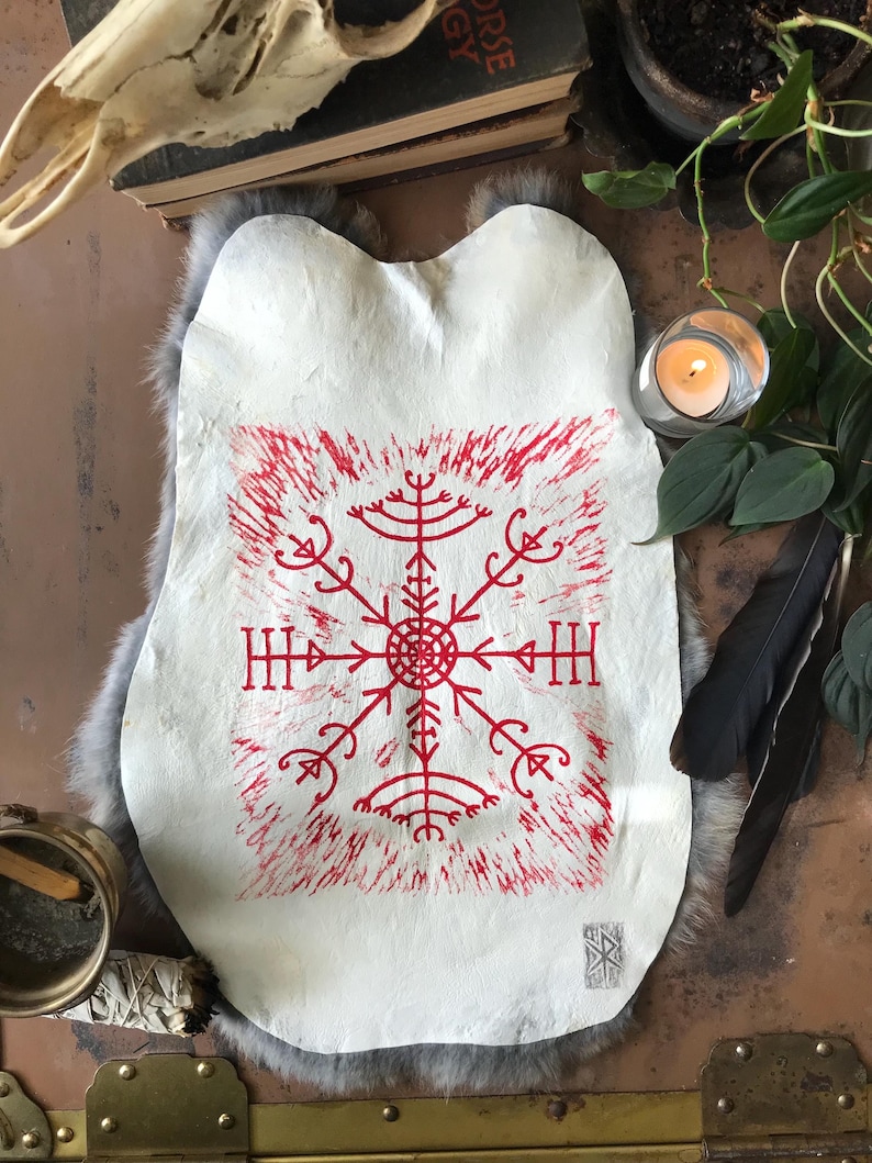 May include: A white, irregularly shaped piece of material with a red, stylized symbol at the center. The symbol is surrounded by a red, textured border. The item is displayed on a gray, furry surface, with a candle and other decorative items in the background.