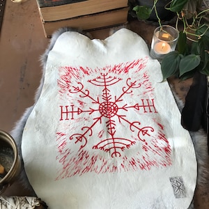 May include: A white leather hide with a red, stylized symbol in the center. The symbol is surrounded by red brushstrokes. The hide is on a gray fur-like surface. A skull, books, and a lit candle are in the background.