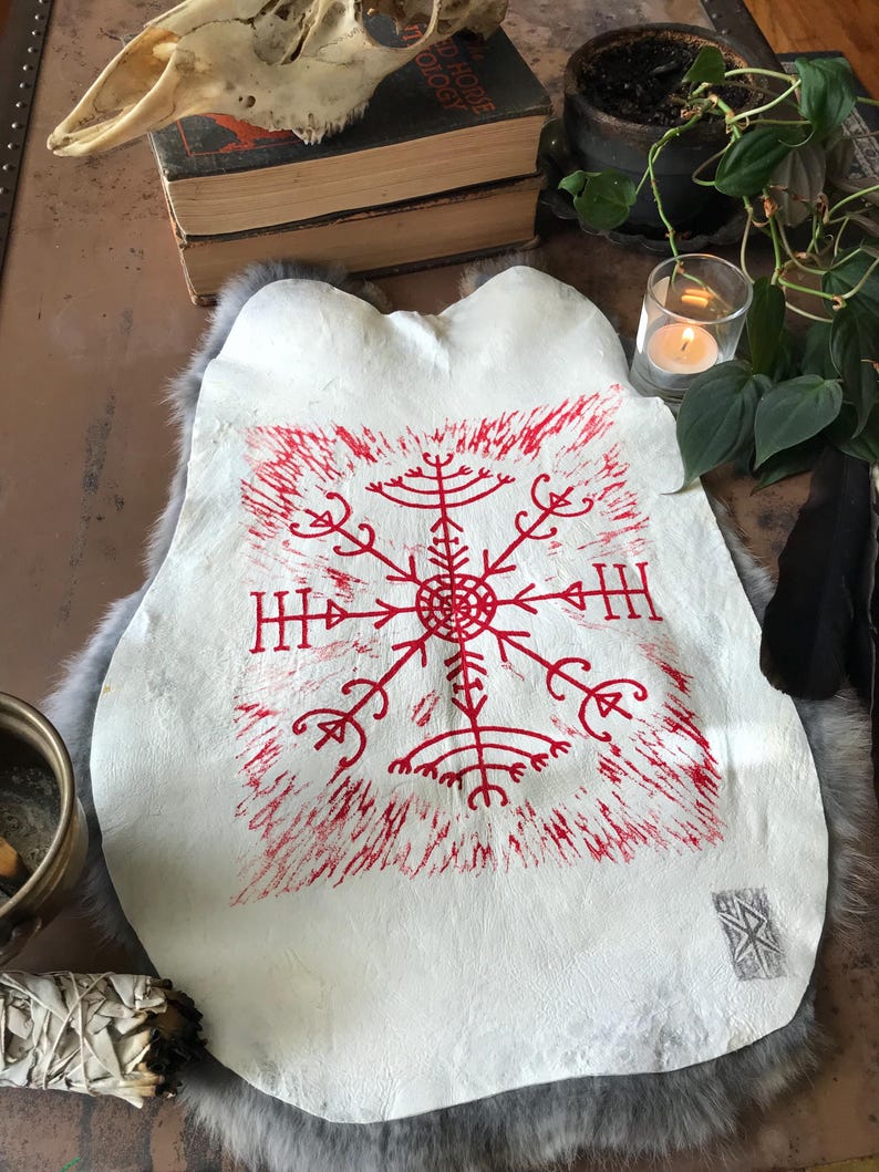 May include: A white leather hide with a red, stylized symbol in the center. The symbol is surrounded by red brushstrokes. The hide is on a gray fur-like surface. A skull, books, and a lit candle are in the background.