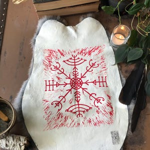 May include: A white leather hide with a red, stylized, geometric design. The design is centered and surrounded by a red, textured border. The hide is resting on a gray, furry surface, with a skull and books in the background.