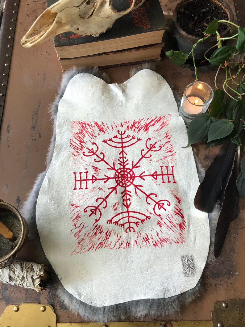 May include: A white leather hide with a red, stylized, geometric design. The design is centered and surrounded by a red, textured border. The hide is resting on a gray, furry surface, with a skull and books in the background.