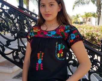 May include: A black embroidered blouse with colorful floral designs. The blouse has short sleeves and a round neckline. The model is wearing the blouse with a black skirt.