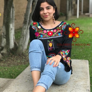 May include: A woman wearing a black embroidered blouse with colorful floral designs, blue jeans, and green sandals. The blouse has a scoop neckline and 3/4 sleeves. The text "COLORES DE MÉXICO MX" is visible on the bottom right corner of the image.