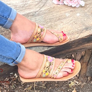 K&ouml;nnte beinhalten: Tanfarbene Ledersandalen mit Blumenstickerei. Die Sandalen haben einen Zehentrenner und einen kleinen Absatz. Die Stickerei ist in Rosa-, Gelb- und Gr&uuml;nt&ouml;nen gehalten.