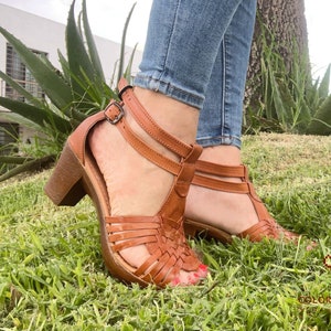 May include: A pair of brown leather sandals with a woven design and a chunky heel. The sandals are worn by a person with blue jeans.