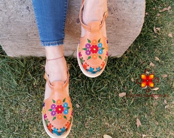 Puede incluir: Un par de sandalias de cuero color canela con bordado floral de colores. Las sandalias tienen una correa a través del pie y una correa de tobillo.