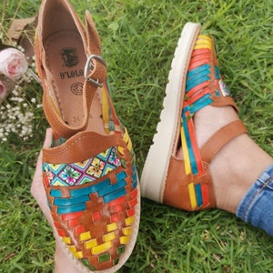 May include: Brown leather sandals with woven colorful straps. The sandals feature a buckle closure and a white sole. The woven design incorporates red, yellow, blue, and green hues. The sandals are displayed on a bed of green grass.