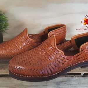 Könnte beinhalten: Ein Paar gewebte Lederschuhe in einem warmen Braunton. Die Schuhe haben ein Slip-on-Design mit offener Ferse und einem gewebten Muster auf der Oberseite. Die Sohlen sind dunkelbraun mit weißen Nähten. Die Schuhe werden auf einer Holzoberfläche präsentiert.