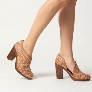 Może przedstawiać: Brązowe skórzane botki z kwiatowym wzorem i masywnym drewnianym obcasem. Botki mają otwarty bok i są noszone z brązową spódnicą. Obcasy mają wysokość około 7,6 cm.