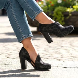 Könnte beinhalten: Schwarze Stiefeletten mit floralem Muster und Blockabsatz. Die Schuhe werden mit blauen Jeans getragen. Der Hintergrund zeigt eine verschwommene Außenszene mit Pflanzen und Blumen.