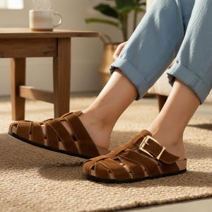 Peut inclure: Sandales en daim marron avec un design de lanières tressées et une fermeture à boucle. Les sandales ont une semelle intérieure en liège et une semelle noire. La personne porte un pantalon bleu et est assise sur un canapé beige.