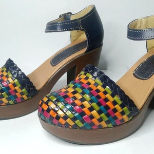 Puede incluir: Un par de sandalias de tacón de cuero tejido con un patrón de cuadros coloridos en tonos de azul, verde, naranja, amarillo, rosa y negro. Las sandalias tienen una correa de tobillo azul marino con cierre de hebilla y una plataforma de madera marrón.