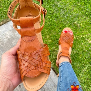 May include: Brown leather sandals with woven straps and a buckle closure. The sandals are on a person's foot and the background is green grass.