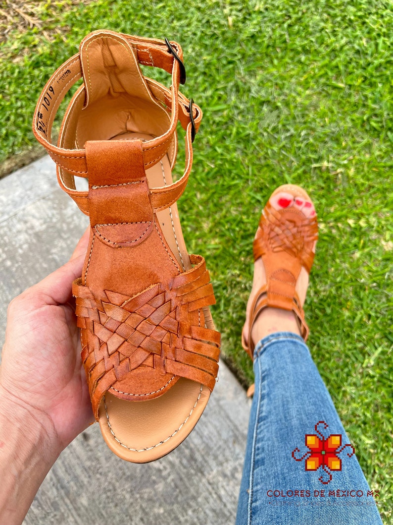 May include: Brown leather sandals with woven straps and a buckle closure. The sandals are on a person's foot and the background is green grass.