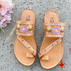 K&ouml;nnte beinhalten: Ein Paar braune Ledersandalen mit Blumenstickerei. Die Sandalen haben eine Zehenschlaufe und einen Riemen, der &uuml;ber den Fu&szlig; geht. Die Stickerei ist in den Farben Rosa, Lila und Gr&uuml;n gehalten.