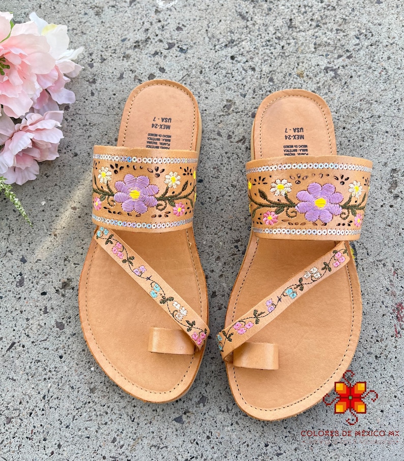 K&ouml;nnte beinhalten: Ein Paar braune Ledersandalen mit Blumenstickerei. Die Sandalen haben eine Zehenschlaufe und einen Riemen, der &uuml;ber den Fu&szlig; geht. Die Stickerei ist in den Farben Rosa, Lila und Gr&uuml;n gehalten.
