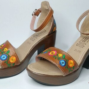 May include: A pair of brown leather sandals with colorful embroidered flowers. The sandals have a chunky heel and an ankle strap.