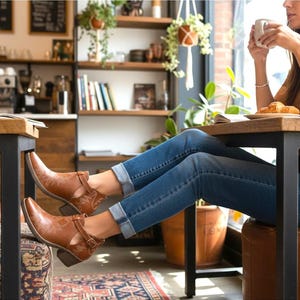 Pu&ograve; includere: Stivaletti in pelle marrone con dettagli traforati e chiusure con fibbia. Gli stivali sono indossati con jeans blu. L'immagine &egrave; scattata in un bar con una persona seduta a un tavolo.