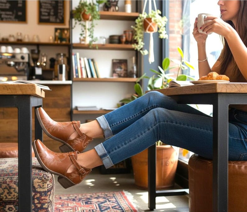 Pu&ograve; includere: Stivaletti in pelle marrone con dettagli traforati e chiusure con fibbia. Gli stivali sono indossati con jeans blu. L'immagine &egrave; scattata in un bar con una persona seduta a un tavolo.