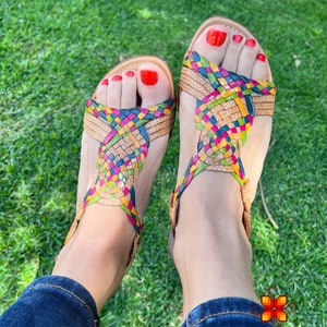 May include: A pair of woven leather sandals with a multi-coloured design. The sandals are being worn on a person's feet, with red nail polish on their toes.