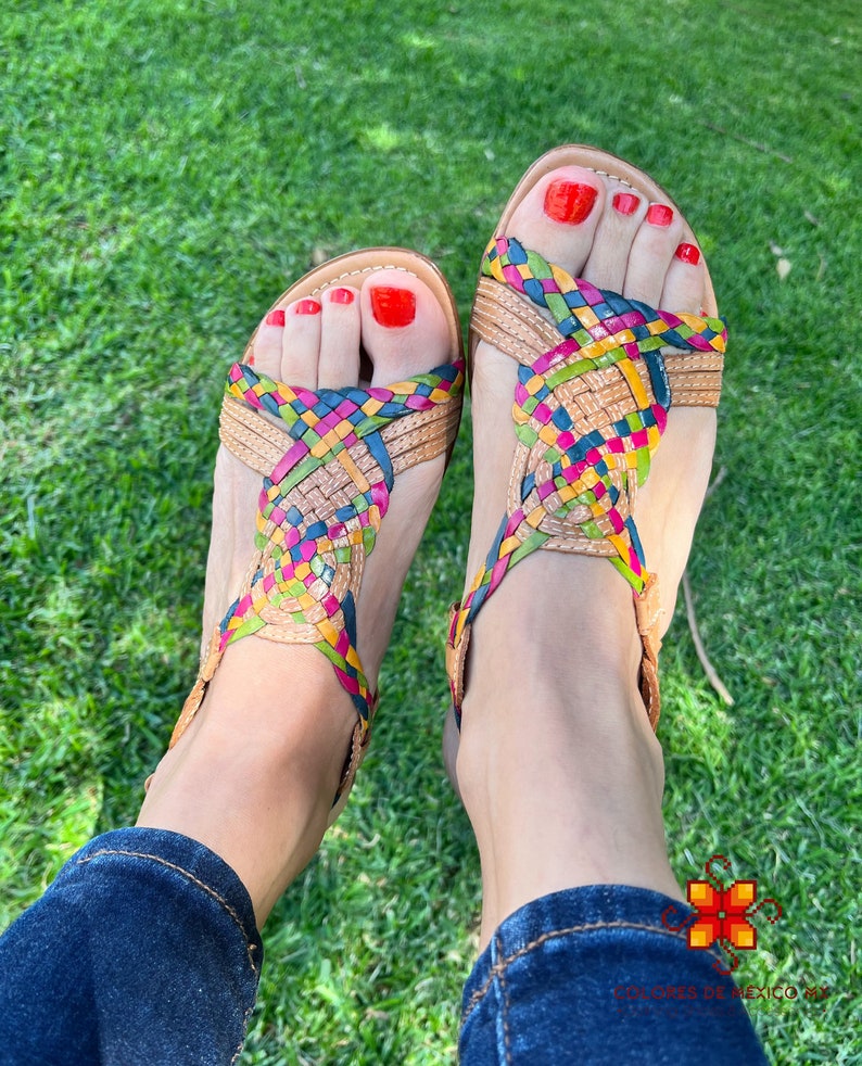 May include: A pair of woven leather sandals with a multi-coloured design. The sandals are being worn on a person's feet, with red nail polish on their toes.