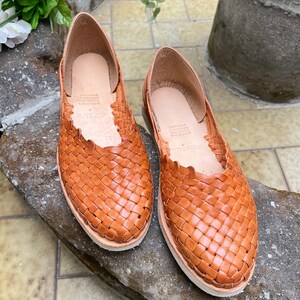 Pu&ograve; includere: Un paio di sandali piatti in pelle intrecciata marroni. I sandali hanno un design semplice e sono realizzati in pelle morbida e flessibile.