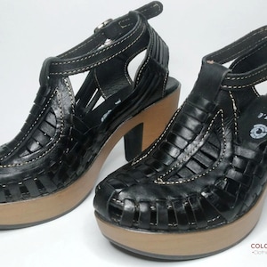 Puede incluir: Un par de sandalias de plataforma de cuero negro con diseño tejido. Las sandalias tienen tacón alto y correa en el tobillo. Las suelas son de color marrón claro. Tacones de unos 10 cm.