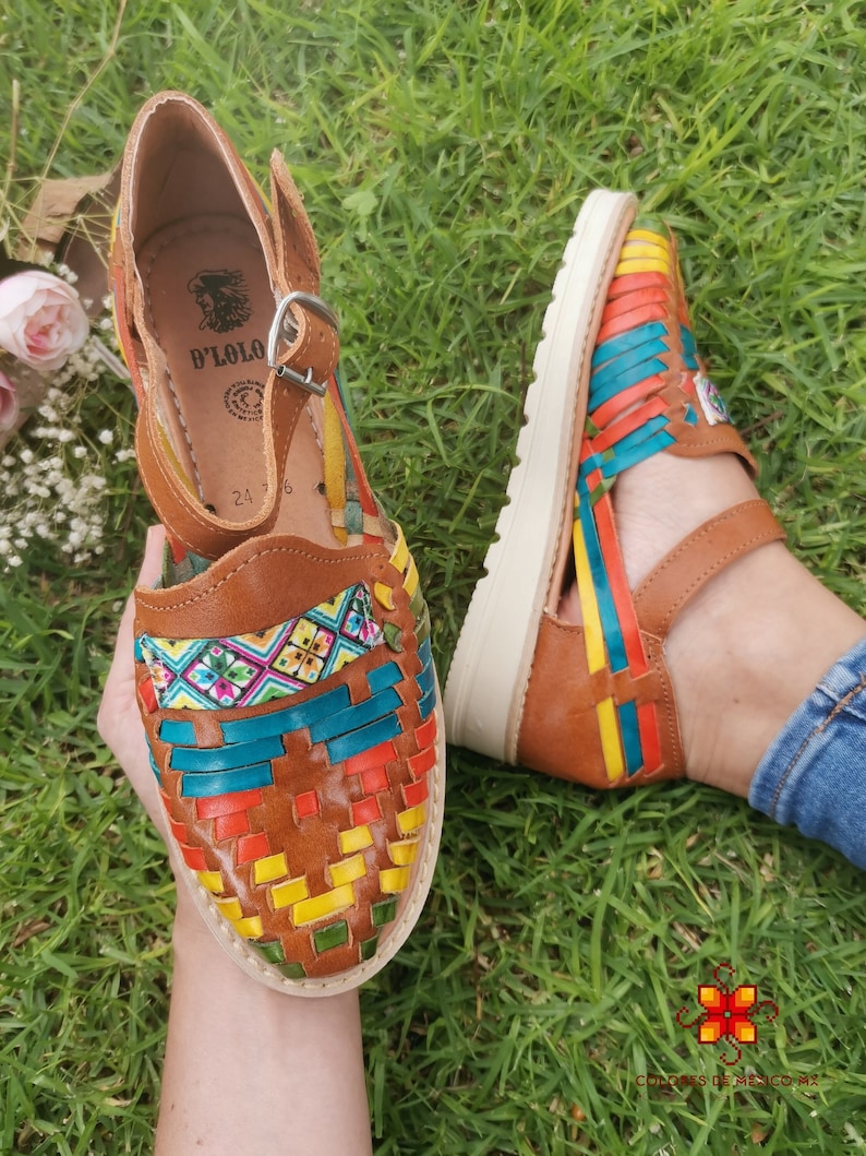 May include: Brown leather sandals with woven colorful straps. The sandals feature a buckle closure and a white sole. The woven design incorporates red, yellow, blue, and green hues. The sandals are displayed on a bed of green grass.
