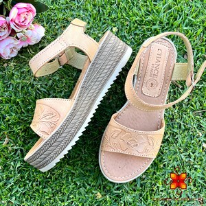 Peut inclure: Une paire de sandales en cuir beige avec un motif floral. Les sandales ont une &eacute;paisse semelle plateforme et une bride &agrave; la cheville avec une fermeture &agrave; boucle. Les sandales sont sur une surface herbeuse verte. Le nom de la marque "CHANCE ESPADRAS" est visible sur la sandale droite.