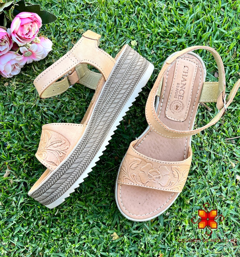 Peut inclure: Une paire de sandales en cuir beige avec un motif floral. Les sandales ont une &eacute;paisse semelle plateforme et une bride &agrave; la cheville avec une fermeture &agrave; boucle. Les sandales sont sur une surface herbeuse verte. Le nom de la marque "CHANCE ESPADRAS" est visible sur la sandale droite.