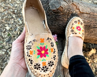 May include: Tan leather slip-on shoes with colorful floral embroidery and cut-out details. The shoes have a jute sole and are shown on a wooden log.
