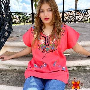 May include: A woman wearing a pink embroidered blouse with floral designs and blue jeans. The blouse has a V-neck and short sleeves. The woman is sitting on a set of stairs in front of a gazebo.