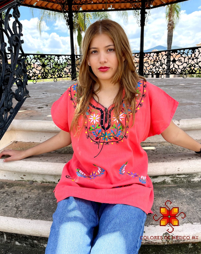 May include: A woman wearing a pink embroidered blouse with floral designs and blue jeans. The blouse has a V-neck and short sleeves. The woman is sitting on a set of stairs in front of a gazebo.