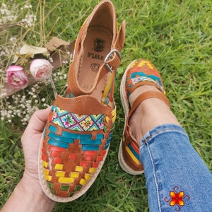 May include: Handmade leather huaraches sandals with colorful woven detailing. The sandals feature a brown leather base with a white sole and a strap with a buckle closure. The woven design incorporates red, yellow, green, and blue accents.