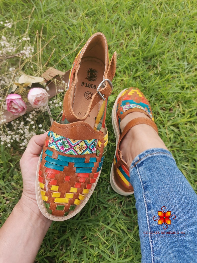 May include: Handmade leather huaraches sandals with colorful woven detailing. The sandals feature a brown leather base with a white sole and a strap with a buckle closure. The woven design incorporates red, yellow, green, and blue accents.