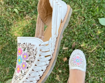 May include: White leather sandals with colorful floral embroidery. The sandals have a woven design and a flat sole.