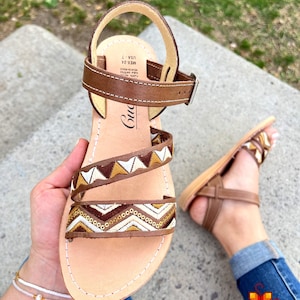 May include: Brown leather sandals with a geometric pattern in white, brown, and gold. The sandals have a buckle closure and are made in Mexico.