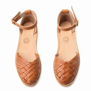 May include: A pair of brown leather sandals with woven straps. The sandals have an ankle strap with a buckle closure.