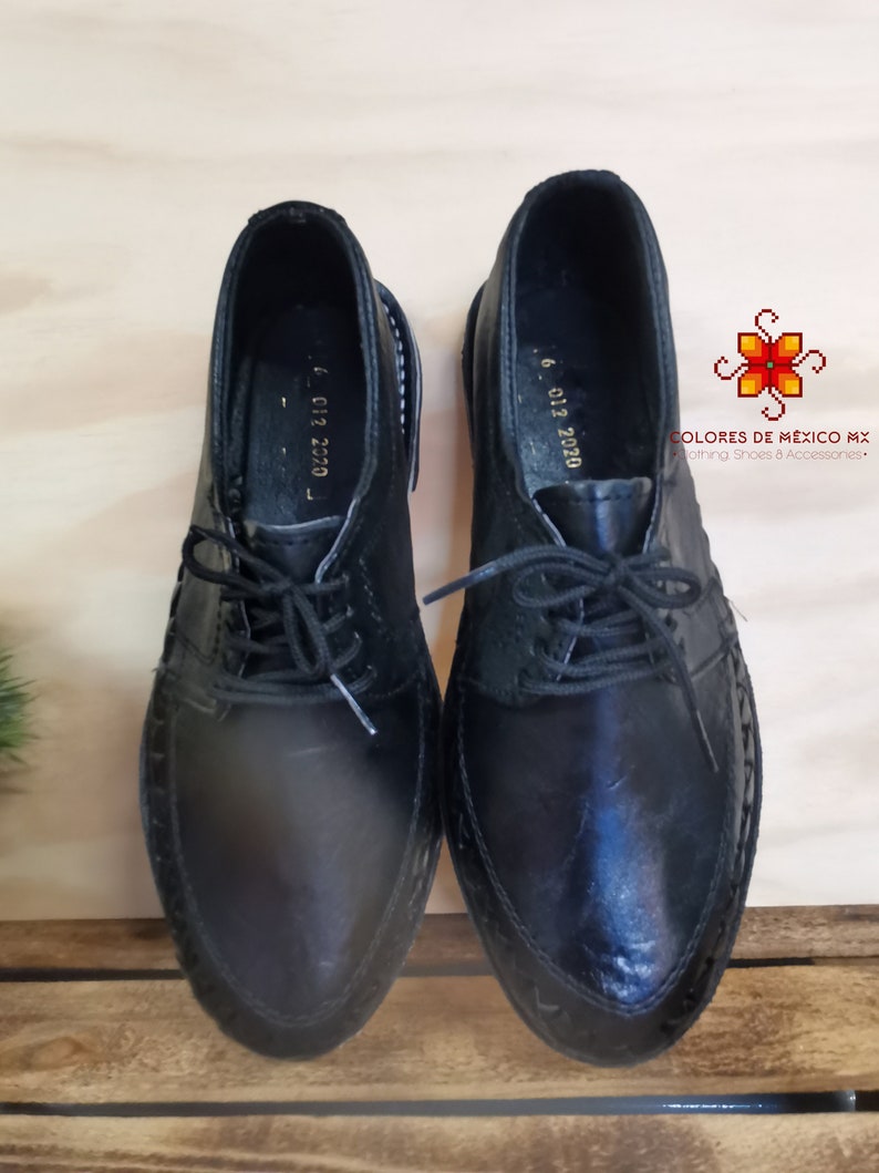 May include: A pair of black leather shoes with laces. The shoes have a pointed toe and a decorative stitch around the edge. The shoes are on a wooden surface.