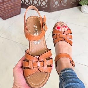 May include: A pair of brown leather sandals with a buckle closure. The sandals have a woven design and a comfortable looking sole.