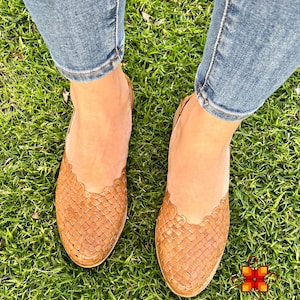 Puede incluir: Un par de sandalias de cuero trenzado marr&oacute;n con un borde festoneado. Las sandalias las lleva una persona de pie sobre hierba verde.