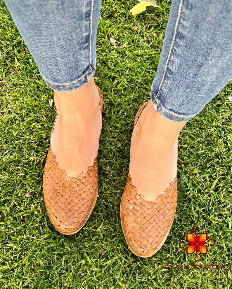 Puede incluir: Un par de sandalias de cuero trenzado marr&oacute;n con un borde festoneado. Las sandalias las lleva una persona de pie sobre hierba verde.