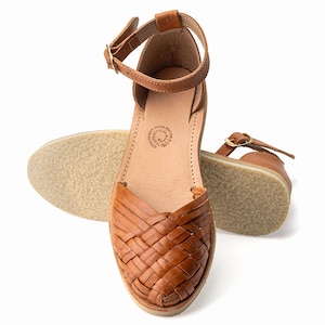 May include: Tan leather sandals with a woven design and an ankle strap. The sandals have a flat sole and are made in Mexico.