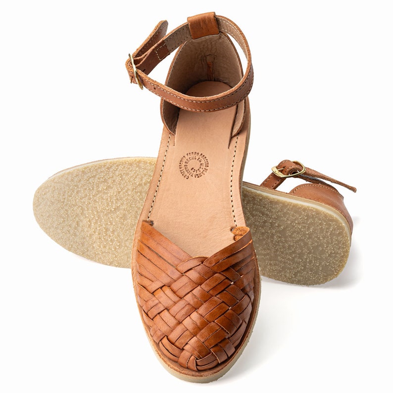May include: Tan leather sandals with a woven design and an ankle strap. The sandals have a flat sole and are made in Mexico.